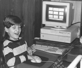 Black and white photo of a boy sitting at a computer desk with his hand on the mouse.