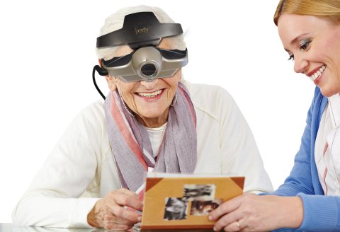 An elderly woman wearing a camera device over her eyes, with the device strapped around her head similar to a headlamp. She is looking at a book with the camera, and a younger woman is beside her.