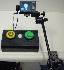 Large rectangular device sitting on a table with a black surface that has a large yellow and green button on the top. Two cables connect it to a digital camera, which is attached to the table mount.