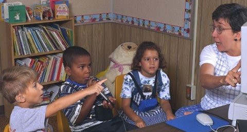 Picture of three young children sitting in front of a woman who is seated at a desk and pointing at a computer screen.