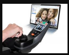 A joystick with a hand of a wheelchair user on it. In front of the user is an iPad being used with Face Time images of people on it.