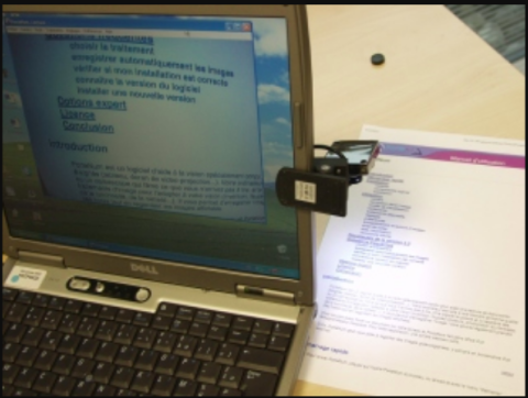 A laptop on a desk with a spring clamp on its screen edge holding an iPhone above a document on the desk.
