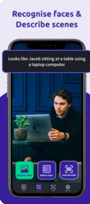 A smartphone's screen featuring a male seated at a table using a laptop. The tagline says, "Recognize faces and describe scenes". The label on the phone reveals what's in the picture: Looks like Jacob sitting at a table using a laptop computer.