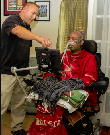A wheelchair user with a technician indicating something on the display screen is shown. The display screen is mounted on the wheelchair with articulating arms.