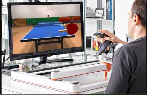 A sideview of a man watching a screen with a virtual ping pong game on it. The man has his wrist supported and strapped in a medical device with his hand clutching an upright handle. There is another person's hand touching the handle also.
