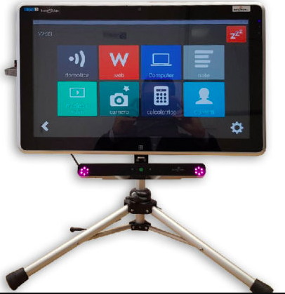 A Tablet that is mounted on a tabletop stand. Attached to its bottom edge is a wide and very slim device that has a small green light in its center and a small circle of white lights at each end. On the screen are large icons like Web, computer, camera, contacts.