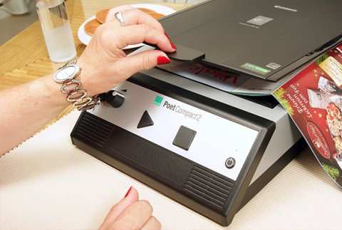 A user's hands in front of a sloped, front panel of a rectangular device that has a large triangle, a large square button and a speaker on its front. One of the user's hands is grasping the scanner lid under which a magazine is on the scanner bed.