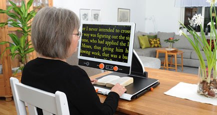 The FHD 17 on a desktop. A woman magnifies a book below the monitor, projecting it in yellow text with a black background. 