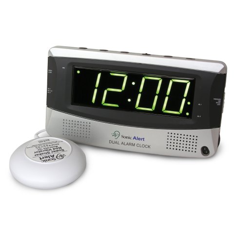 A rectangular, silver, alarm clock with large led display and two speakers on either bottom side of the front face; control buttons on the top face; and, a small, wired, domed shaker device to the alarm clock.