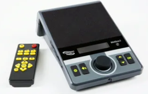 A rectangular and slanted control box divided into a black speaker on the upper half and a lower grey panel with 5 large keys and a dial. Next to this box is a black remote with bright yellow keys and a red power button.