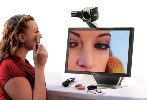 A woman is applying makeup and using the magnifier as a mirror, with the monitor displaying a zoomed-in video feed of her face.