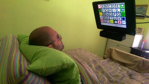 A man lying in bed controlling CiaoMundo with his eye movement. A monitor is positioned on a tall stand over his bed so the user can look at it comfortably.
