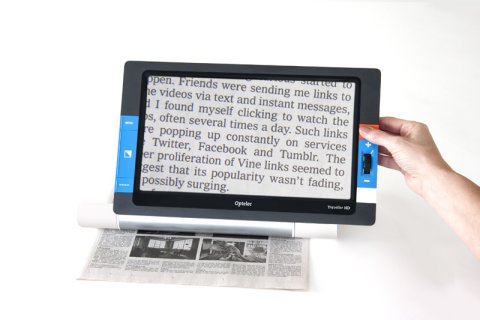 Rectangular tablet with blue control buttons along the edges. A hand holding the tablet above a newspaper and a digital version of a magnified newspaper excerpt displayed on screen.