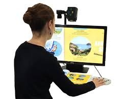 A woman sits in front of a monitor with a magnifier mounted at the top with controls to zoom and a light attached. 