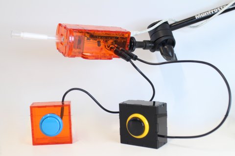 A rectangular orange plastic device with a clear plastic point connected to a flexible arm. Two small square devices are attached with round buttons.