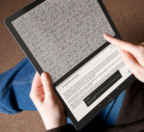 A tablet held in two hands and shows a semi-full screen of braille content, with a small portion below showing the printed version.