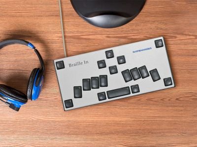 A silver Braille keyboard with black keys placed on top of a wooden surface next to a pair of blue headphones. 
