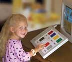 A young girl with blonde hair sits at a computer and points to a board with several different colored squares.