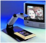 A small camera mounted on an angled neck and connected to a platform base, with space for the user to place print materials on top of and position underneath the camera for magnifying. Next to the camera is a small TV displaying the magnified content (a family photo).