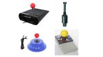Various models of joystick switches. They resemble standard wheelchair joysticks, with long, thin switches with either a small knob or ball on top for the user to grasp and push/pull the switch. One model has a large yellow ball grip; two have smaller red ball grips. One model has a flat black knob or button grip. The joysticks are embedded in small control devices roughly the size of a wheelchair joystick box. These are either rectangular or circular in shape. One is blue; two are black; one is grey.