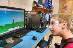 The side-view of a young boy sitting in front of a computer with a colorful screen image of people standing in the grass. The user has electrodes on his temple and forehead.