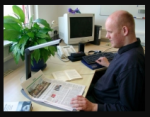 A man is sitting at a desk with his left hand on the bottom edge of a paper and his right on a keyboard that is sitting on top of a braille display. The paper is under a camera for OCR.