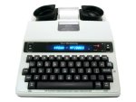 A TTY device resembling an electric typewriter. The keys and display panel are black, while the device body is off-white. 