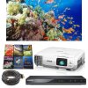 A coral reef scene with brilliant blue and orange colors, a 3x3 grid of colorful DVD covers, a white rectangular projector with a built-in speaker on the front left and a projector lamp/eye on the right, a thin black rectangular DVD player, and a large coil of black wire ending in a USB.